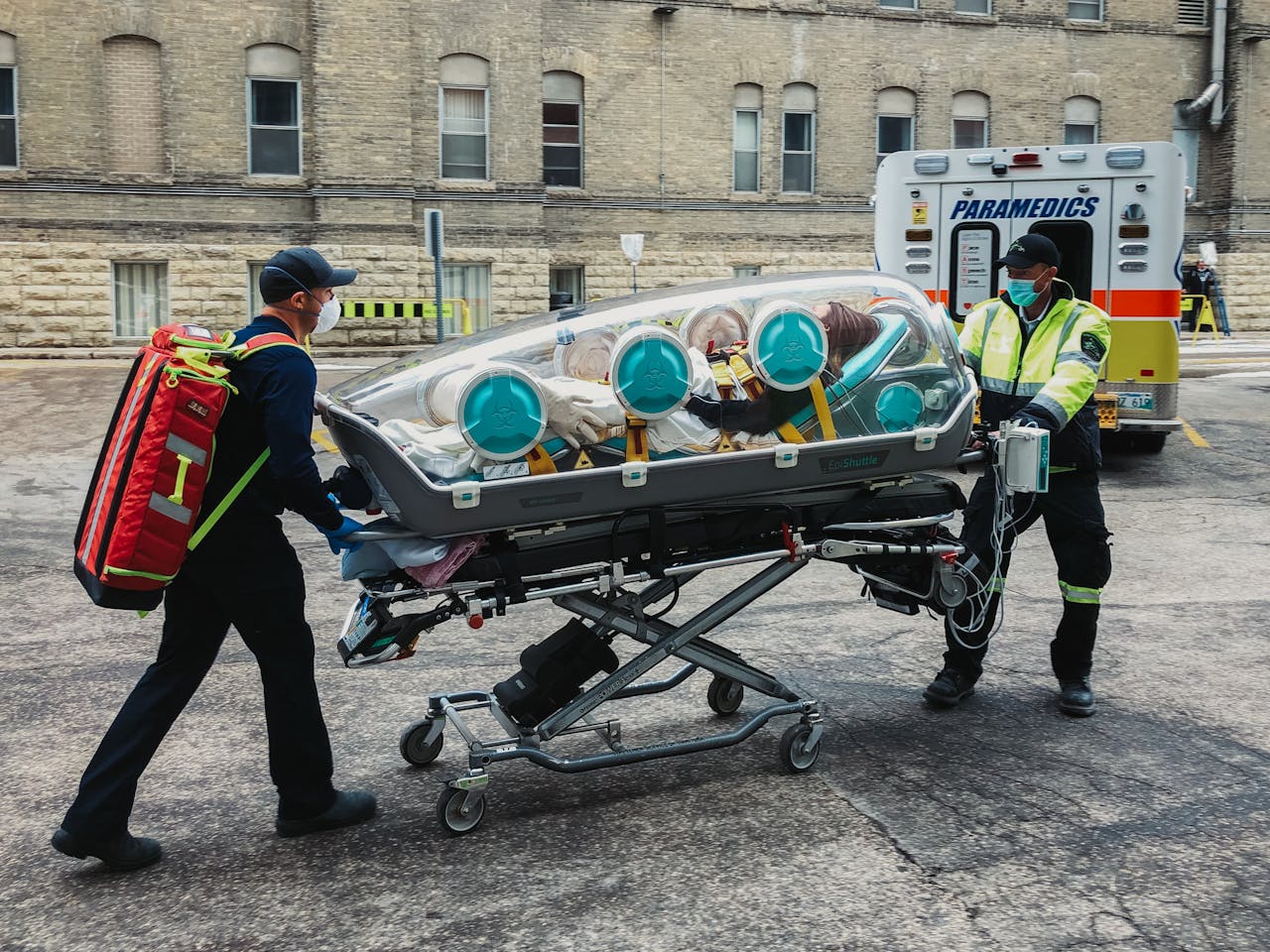 Emergency paramedics transport a patient in an isolation pod outside a medical facility.